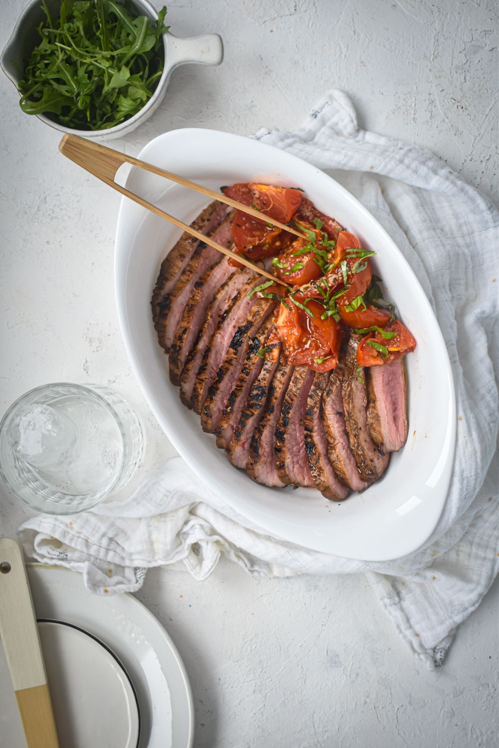 Flank steak with balsamic marinade and roasted tomatoes on a white plate with greens on the side.