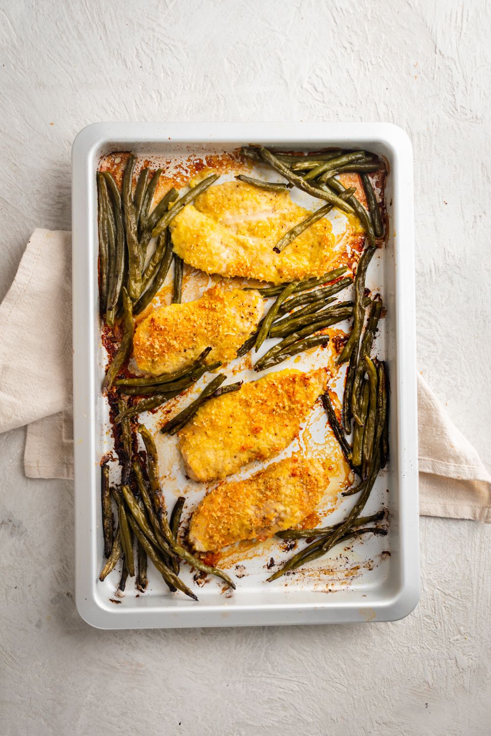 Lemon pepper breaded chicken cutlets and roasted green beans on a white sheet pan.