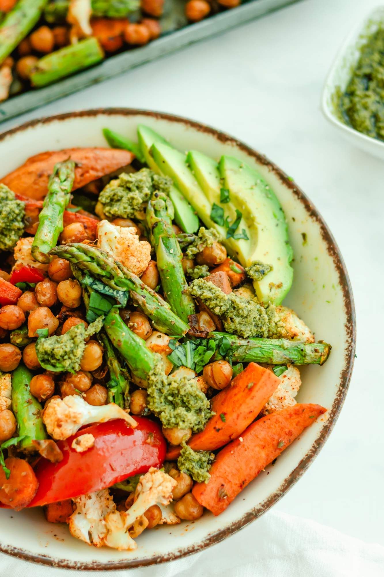 Roasted vegetables and chickpeas including sweet potatoes, carrots, asparagus, cauliflower, red peppers, and chickpeas in a bowl with sliced avocado and pesto.