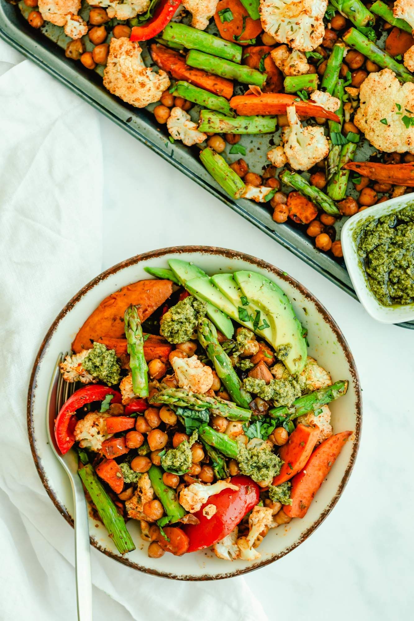 Roasted chickpeas with sweet potatoes, cauliflower, red peppers, asparagus, and avocado in a bowl and on a sheet pan.