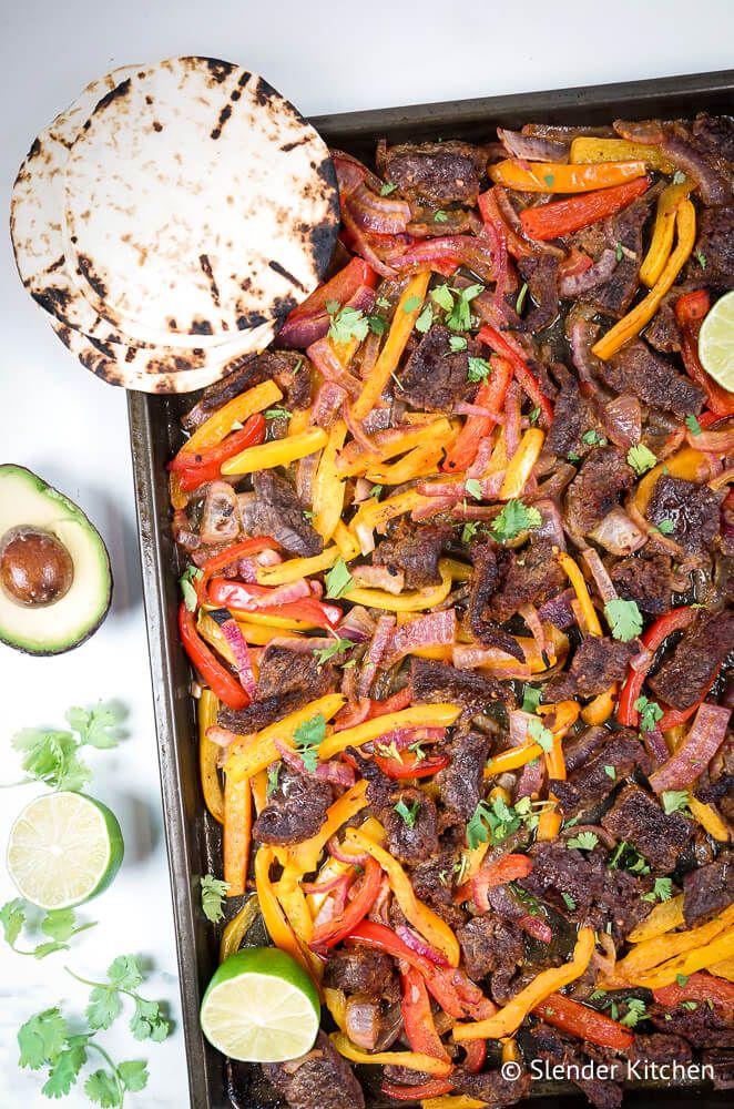 Steak fajitas in a sheet pan with bell peppers, red onions, flour tortillas, cilantro, and limes.