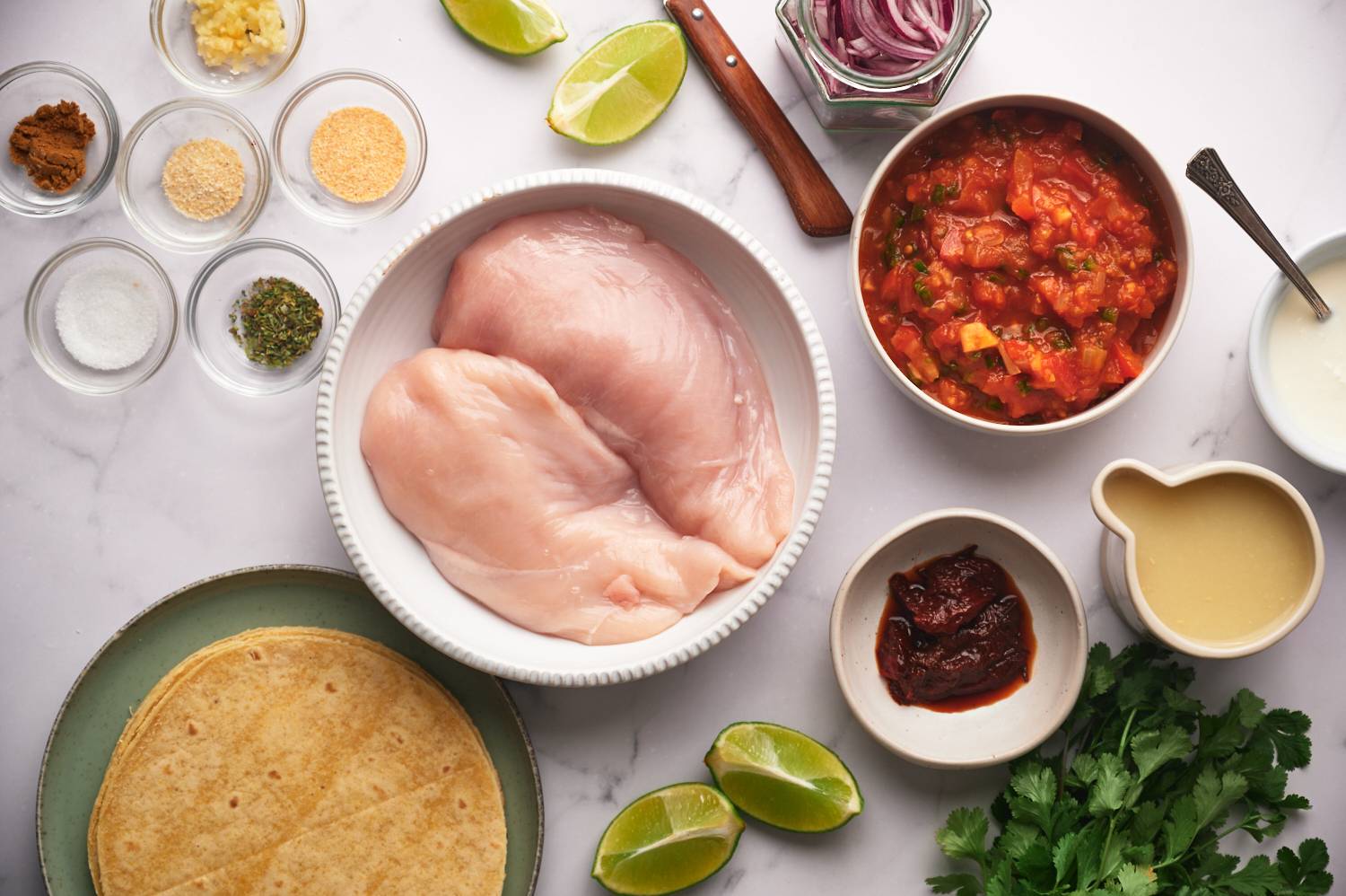 Ingredients for shredded chicken tacos including chicken breast, tomatoes, chipotle, lime juice, spices, and corn tortillas.