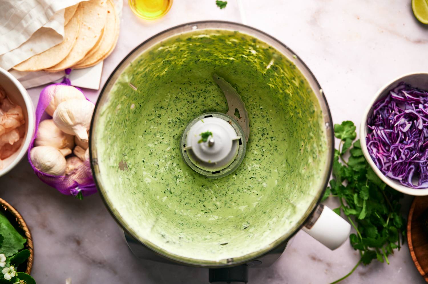 Cilantro lime shrimp taco sauce in a food processor with garlic, cabbage, and tortillas on the side.