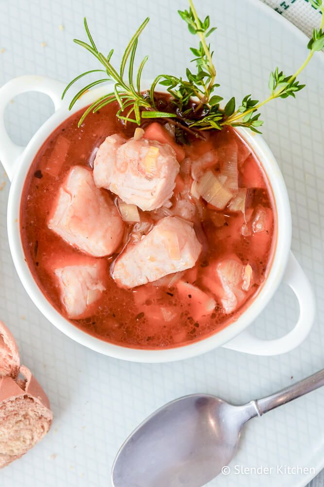 Fish stew in white bowl with red snapper, veggies, tomato broth, and fresh herbs with bread on the side.