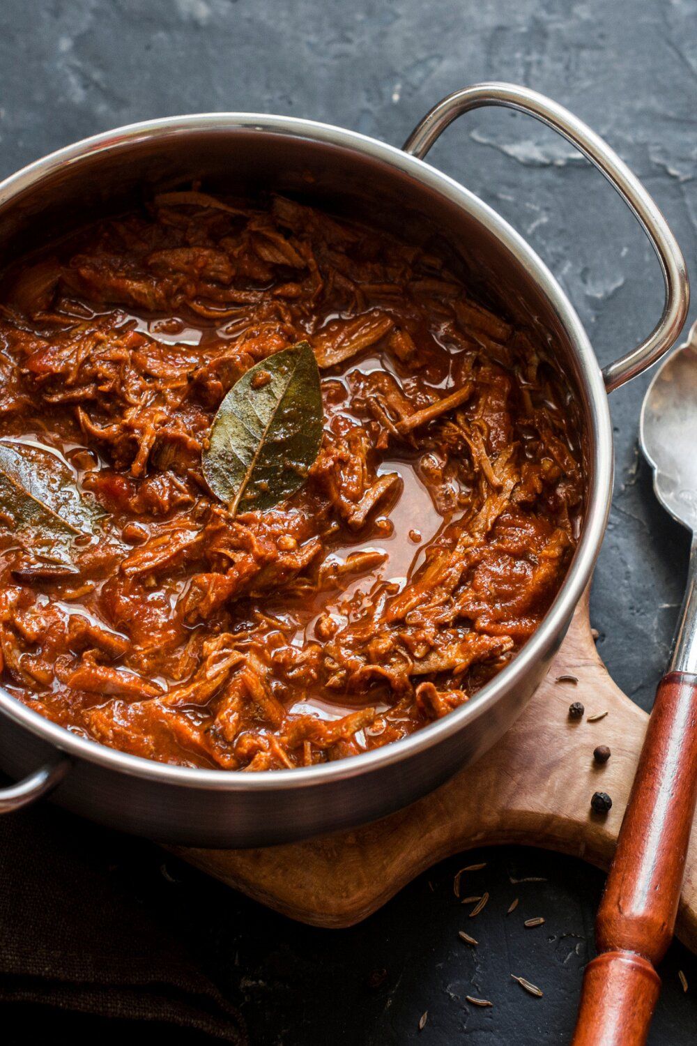 Crockpot beef ragu in a large pan with a bay leaf and spoon on the side.