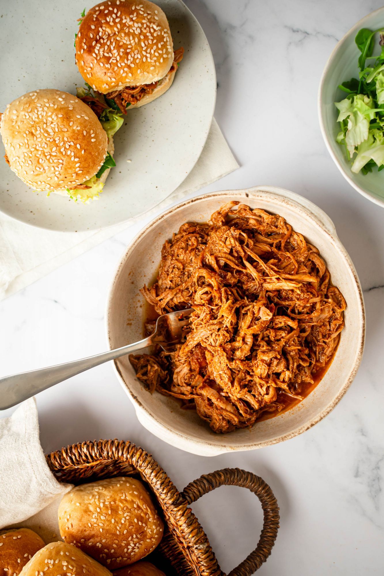 Crockpot spicy pulled pork in a bowl with spicy barbecue sauce with sandwiches on the side.