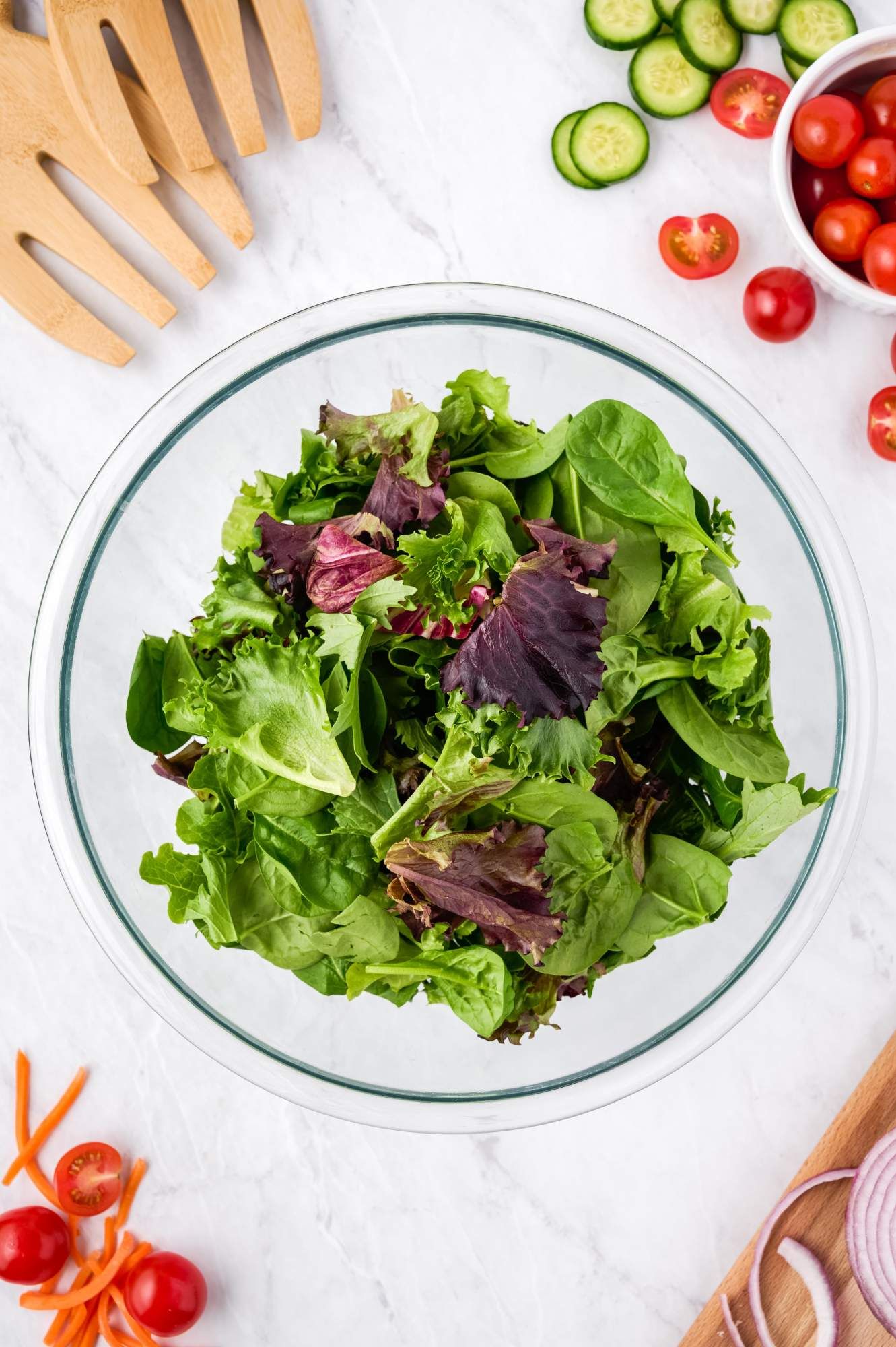 Spring mix with spinach and lettuces in a bowl with tomatoes, cucumbers, and salad tongs on the side.