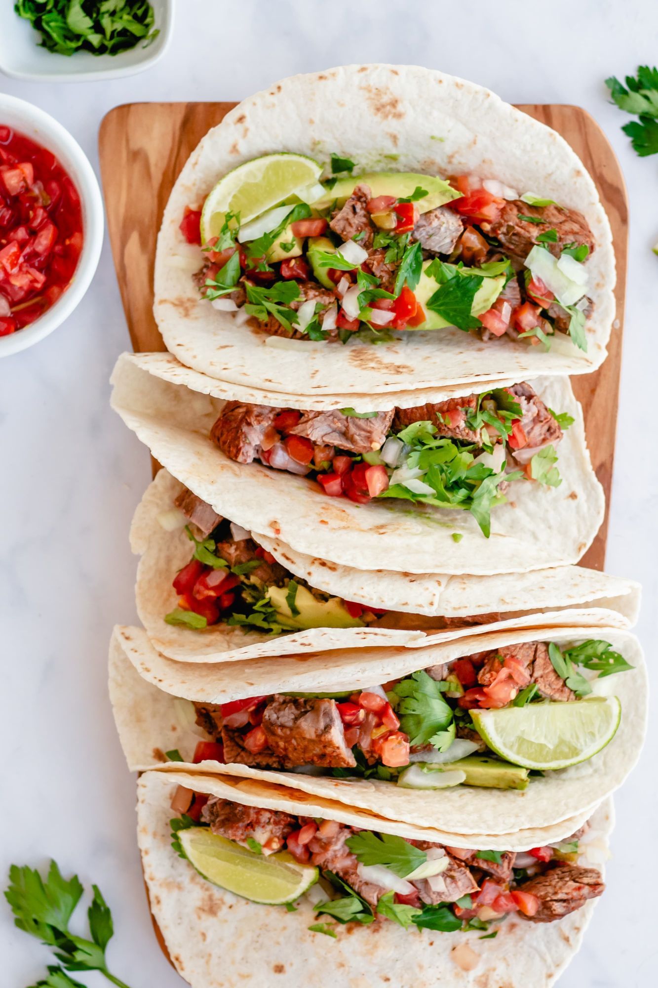 Beef tacos with tender steak, pico de gallo, avocado, and lime wedges in flour tortillas on a cutting board.