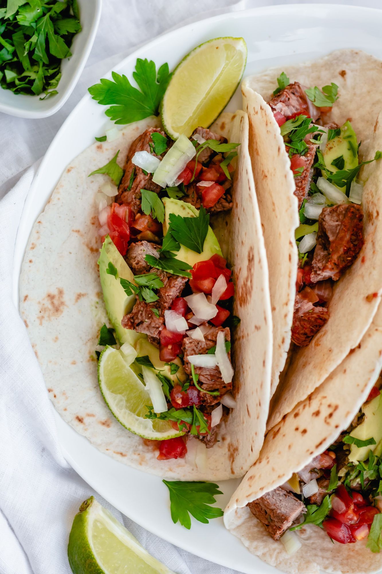 Tender steak tacos with salsa, avocado, cilantro, onion, and tomatoes in a tortilla with limes.