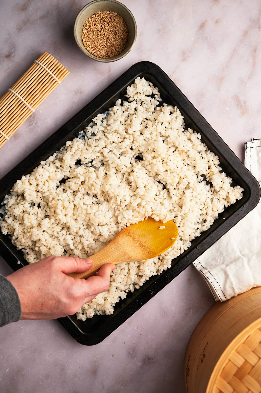 Sushi rice on a baking sheet being stirred with rice vinegar, salt, and sugar.