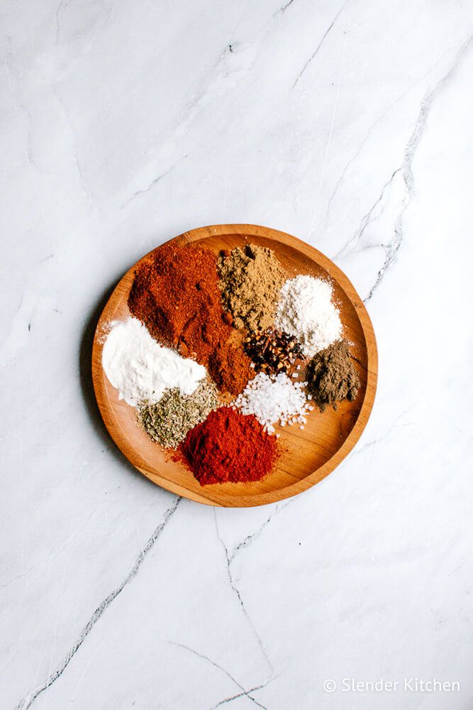 Taco seasoning ingredients including chili powder, paprika, and cumin on a wooden plate with a spoon.