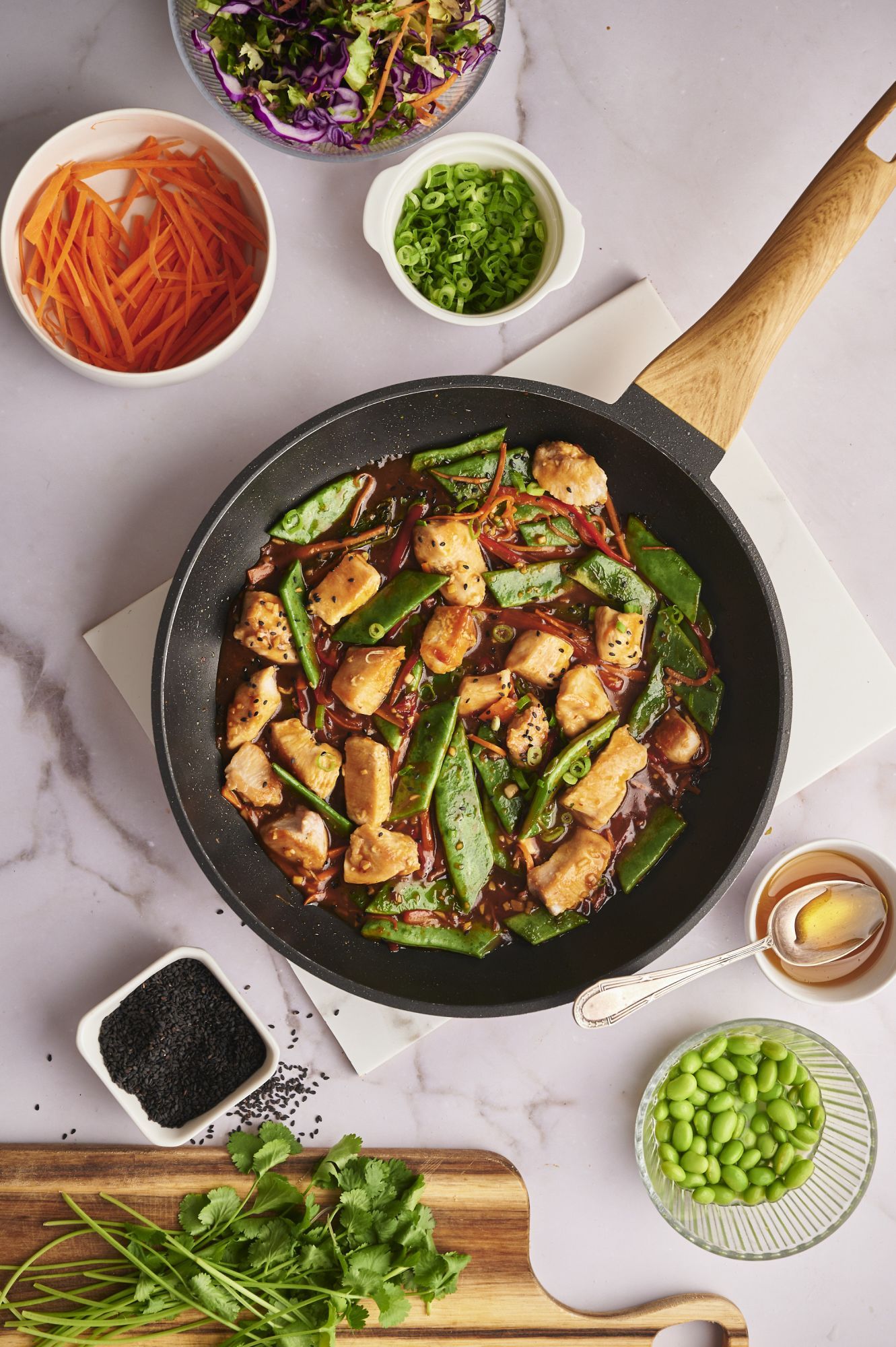 Teriyaki chicken and vegetables in a skillet with vegetables, herbs, and sesame seeds on the side. 