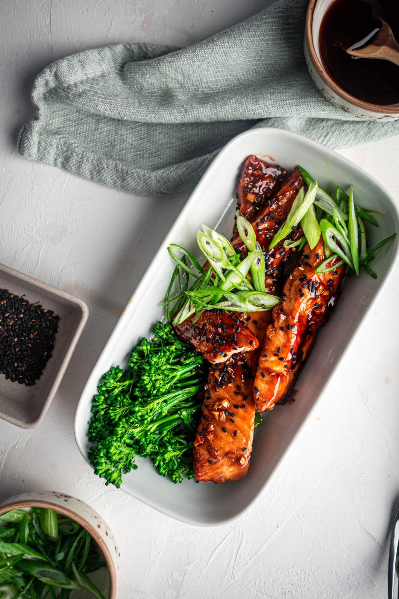 Teriyaki baked salmon on a plate with broccoli, green onions, and sesame seeds with a blue napkin.