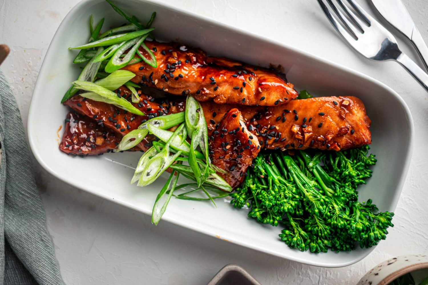 Salmon with teriyaki sauce, sesame seeds, green onions, and broccoli on a white plate with a fork.