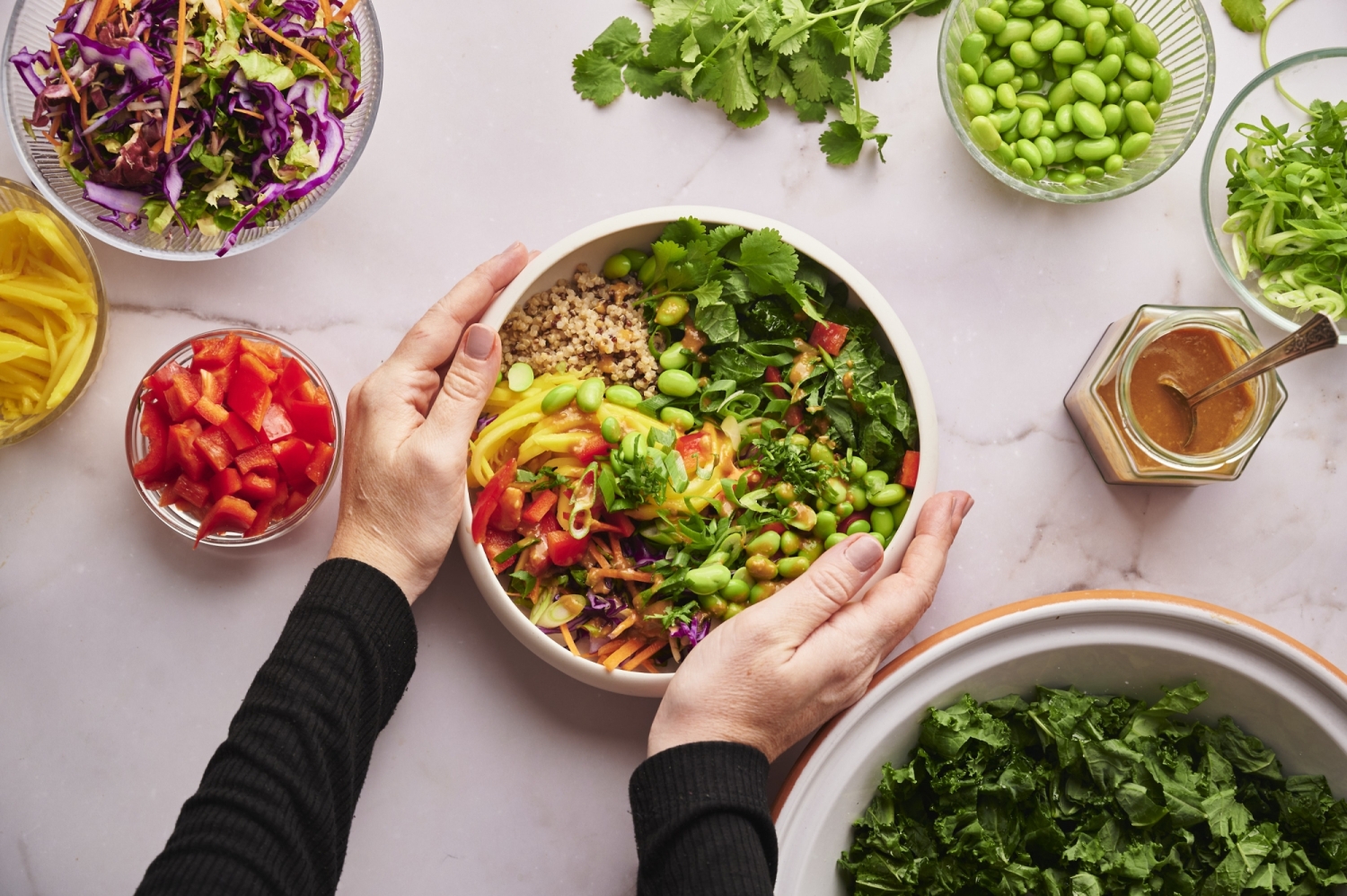 Thai buddha bowl with quinoa, fresh vegetables, edamame, cilantro, and peanut sauce in a bowl with two hands on the side.