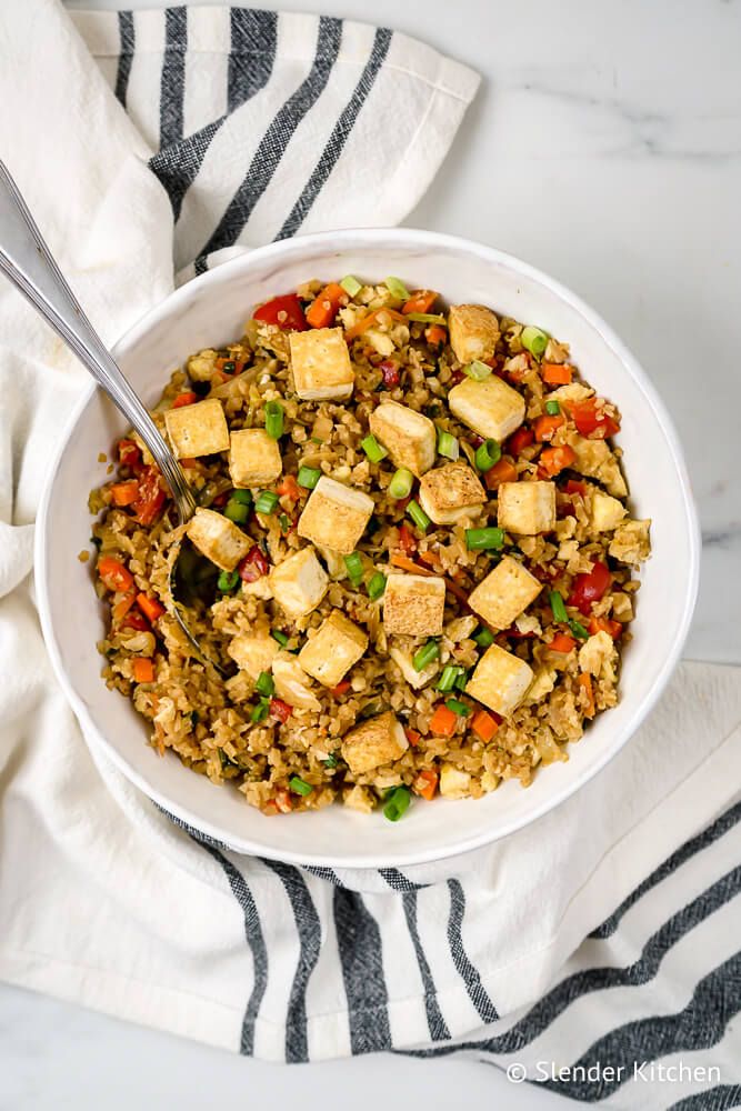 Fried rice with tofu and cauliflower rice in a bowl with vegetables, green onions, and a spoon.