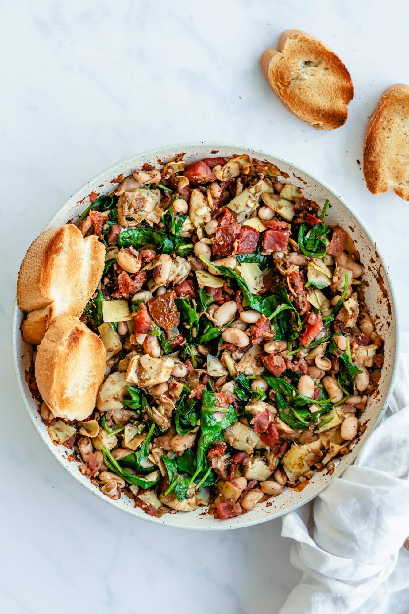 Cannellini beans in a skillet with spinach, artichokes, garlic, onion, tomatoes, and sundried tomatoes.