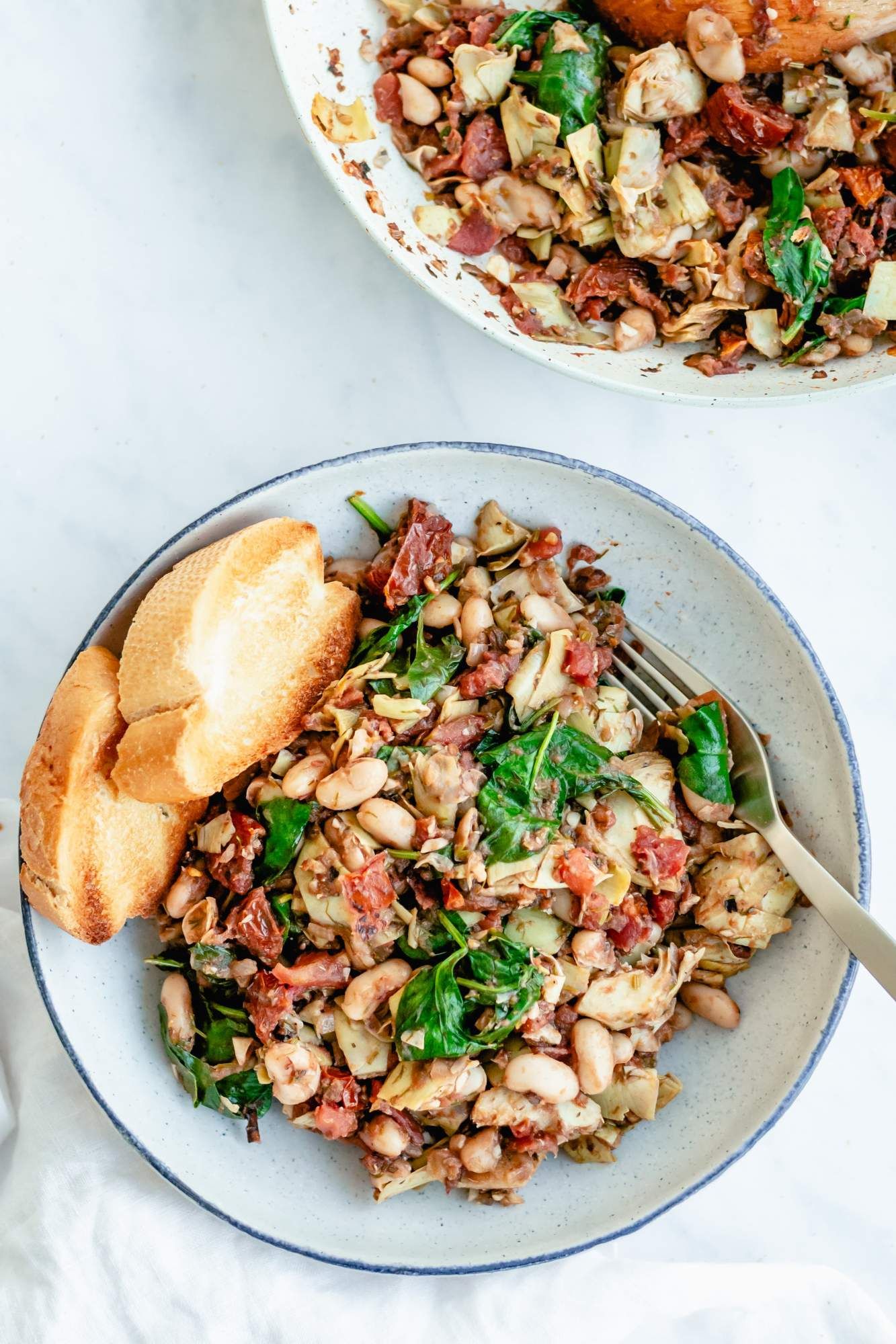 White beans, tomatoes, spinach, and artichoke hearts cooked together and served in a bowl with toast.