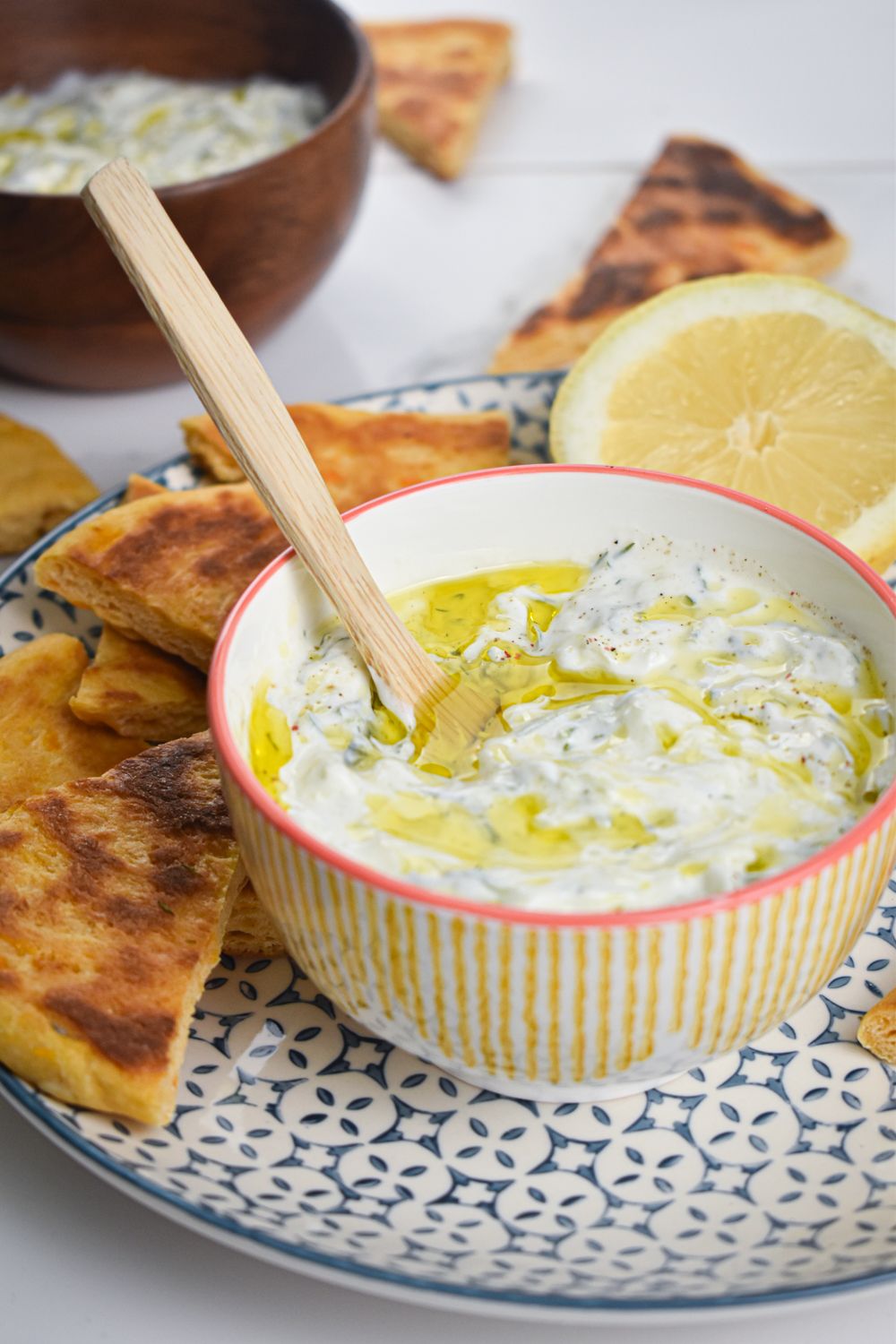 Easy tzatziki sauce in a yellow bowl with a wooden spoon, fresh lemon, and pita wedges.