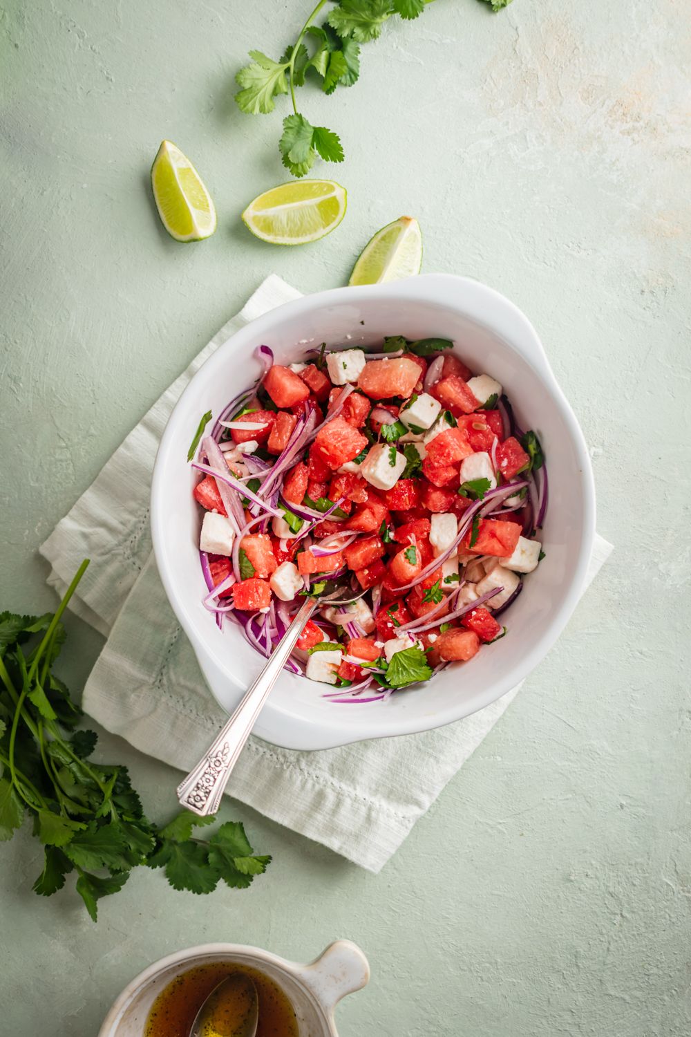 Watermelon salad with feta cheese, red onions, cilantro, and lime juice in a white bowl with dressing on the side.