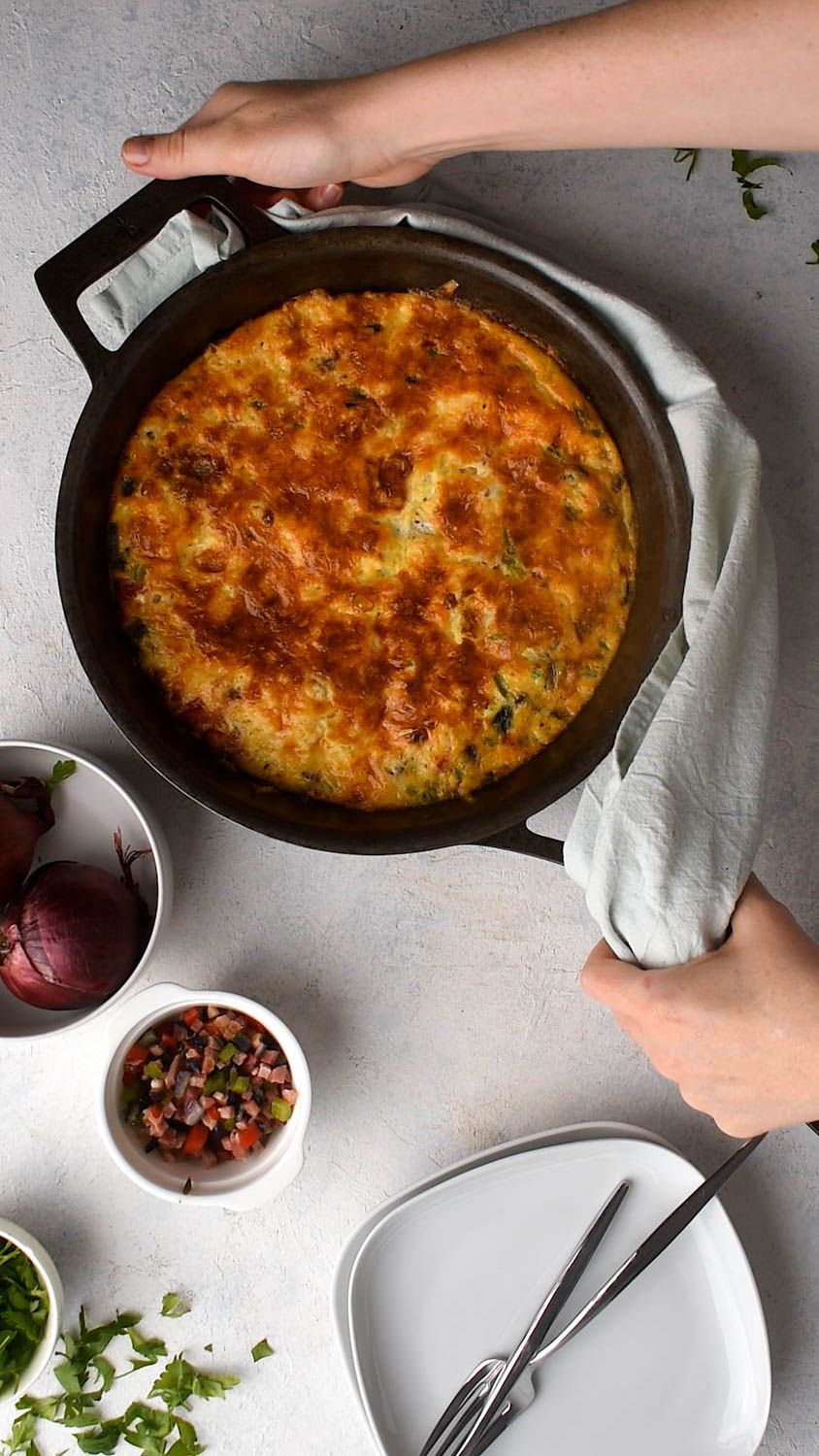 Cast iron skillet with a Western frittata cooked with cheddar cheese, lean ham, peppers, and onions being held by two hands.