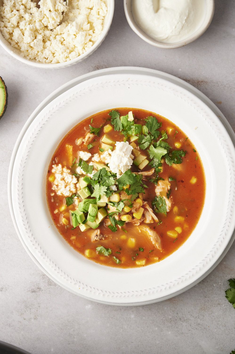 Bowl of Mexican chicken soup with shredded chicken, corn, cilantro, and avocado with sour cream and cheese on the side. 