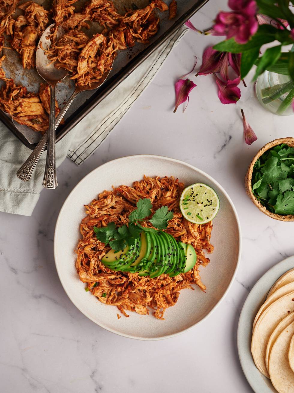 Bowl of Mexican shredded chicken served with cilantro, avocado, lime, and tortillas.