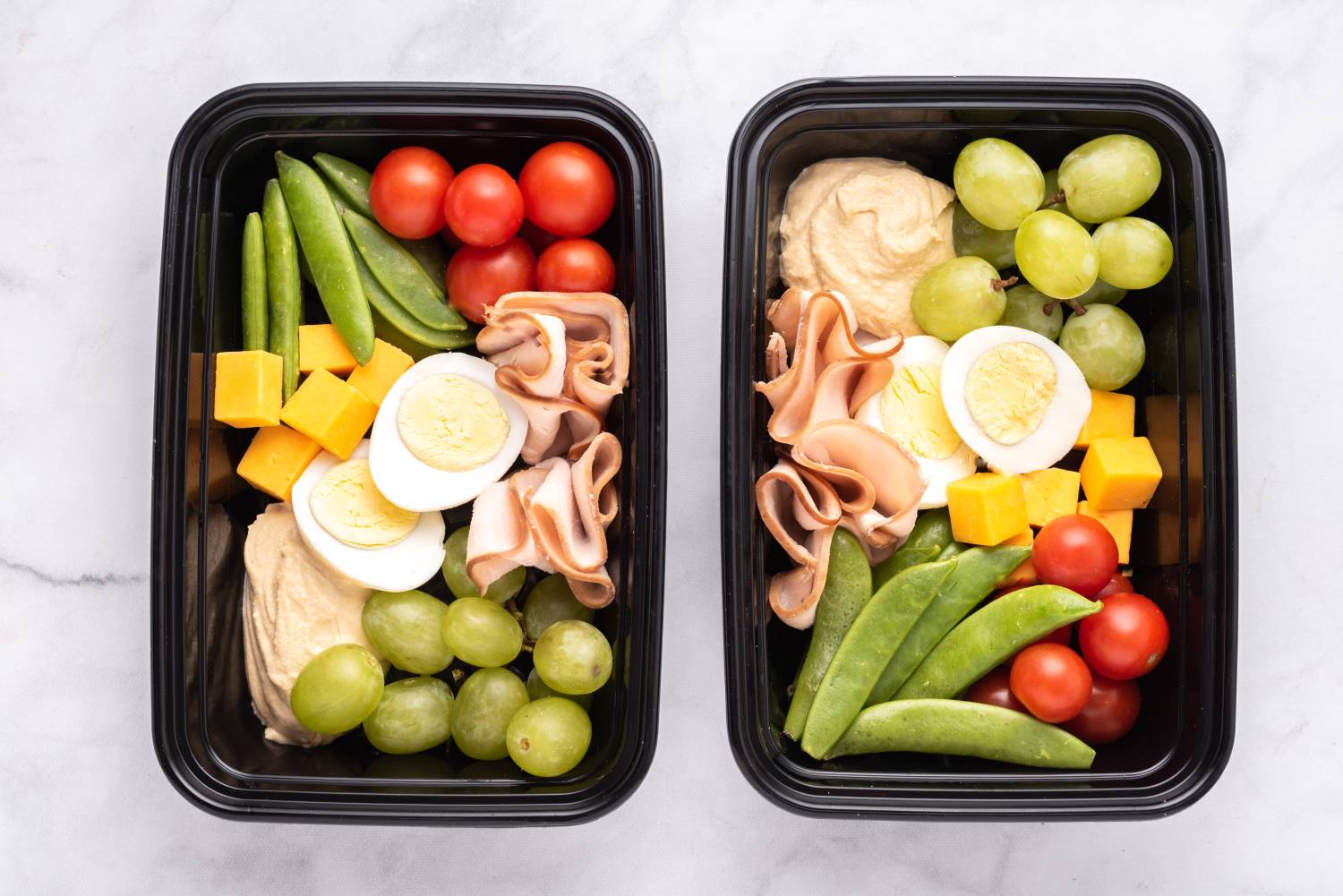 Copycat protein bistro box with grapes, boiled egg, cheese, hummus, turkey, and vegetables in a portable plastic container.