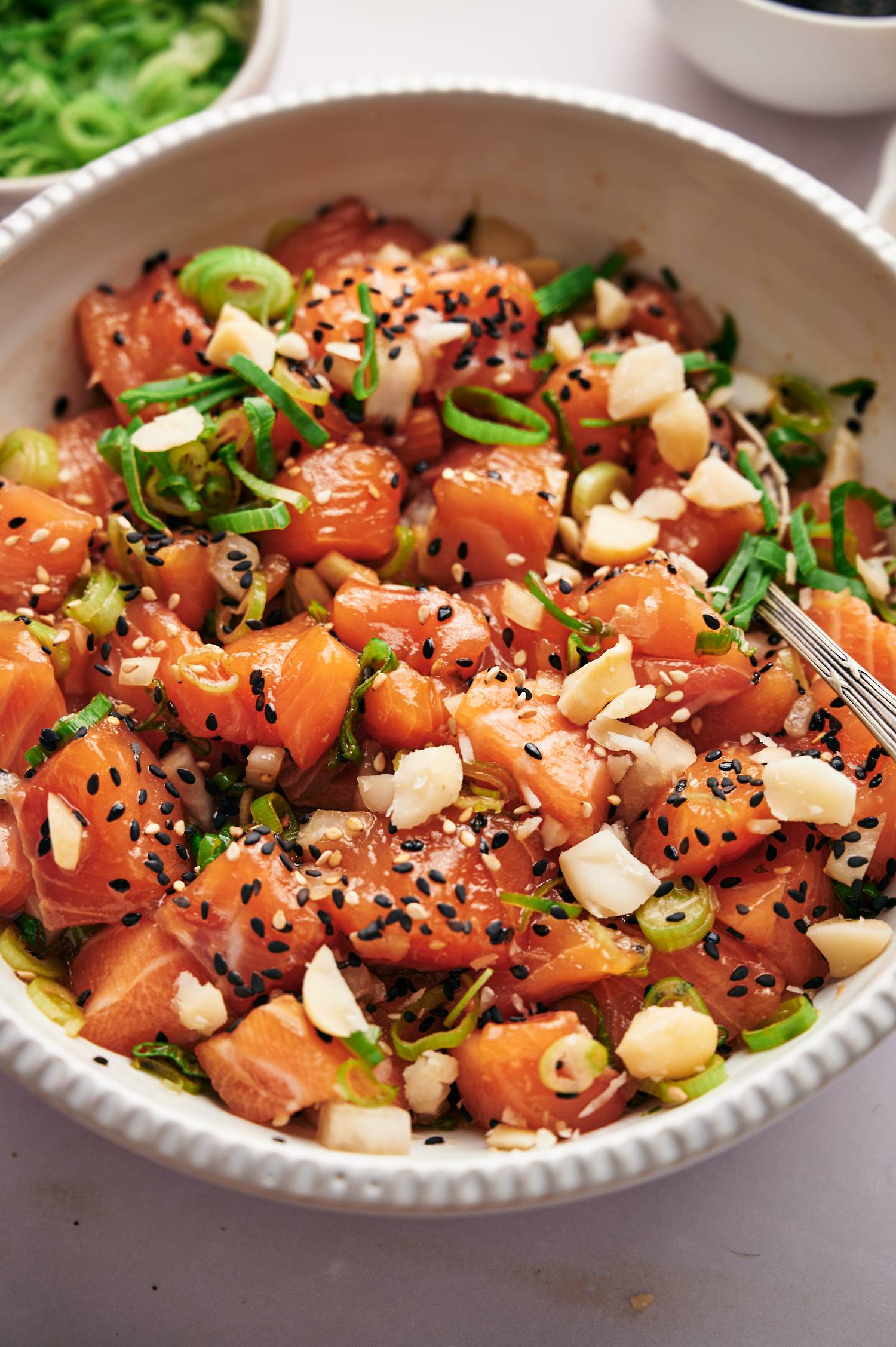 Raw salmon poke with soys sauce, green onions, sesame seeds, and macadamia nuts in a white bowl.