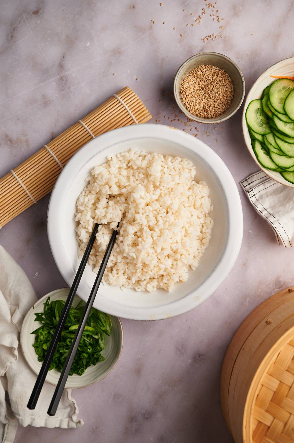 Cooked sushi rice with vegetables, sesame seeds, and green onions on the side with chopsticks.