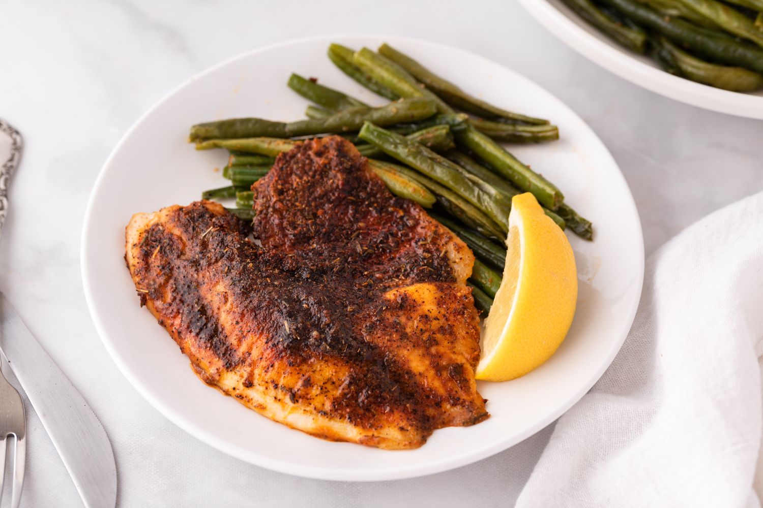 Baked blackened tilapia in a baking dish with lemon slices and blackening seasoning.