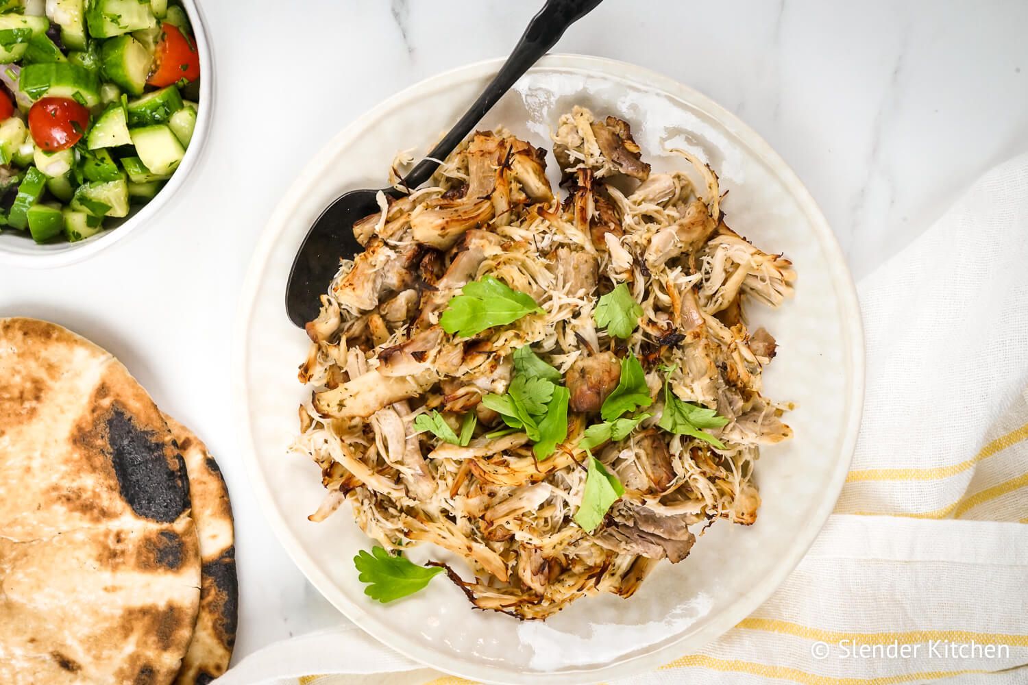 Greek chicken shredded on a plate with oregano, parsley, lemon, pita bread, and cucumber tomato salad.