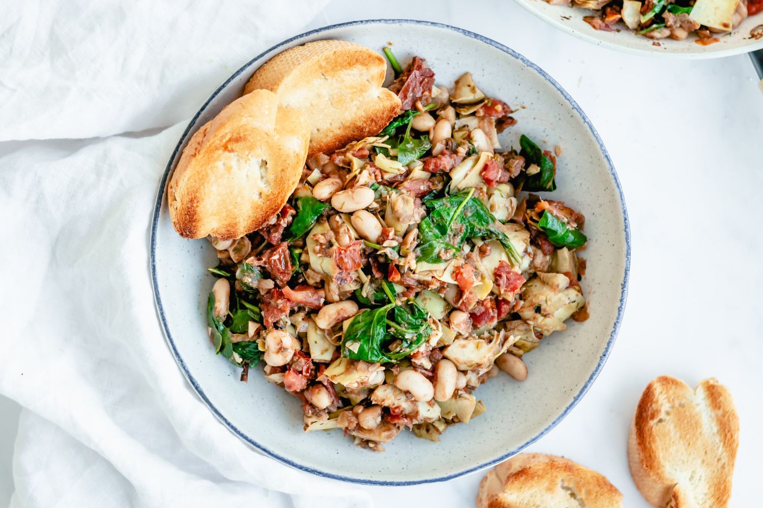 Tuscan white bean skillet with spinach, tomatoes, artichokes, onions, and garlic in a bowl with toast.