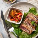 Grilled tuna with soys sauce and sesame seeds on a plate with green onions, lettuce, and cooked peppers.