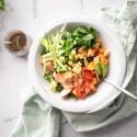 Salmon kale salad with sweet potatoes, avocado, apples, and a maple dijon dressing in a white bowl.