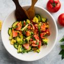 Tomato cucumber salad with corn, avocado, and fresh basil in a bowl with wooden salad tongs.