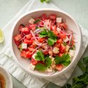 Watermelon and feta salad with cilantro and served with lime vinaigrette in a white bowl with fresh limes on the side.