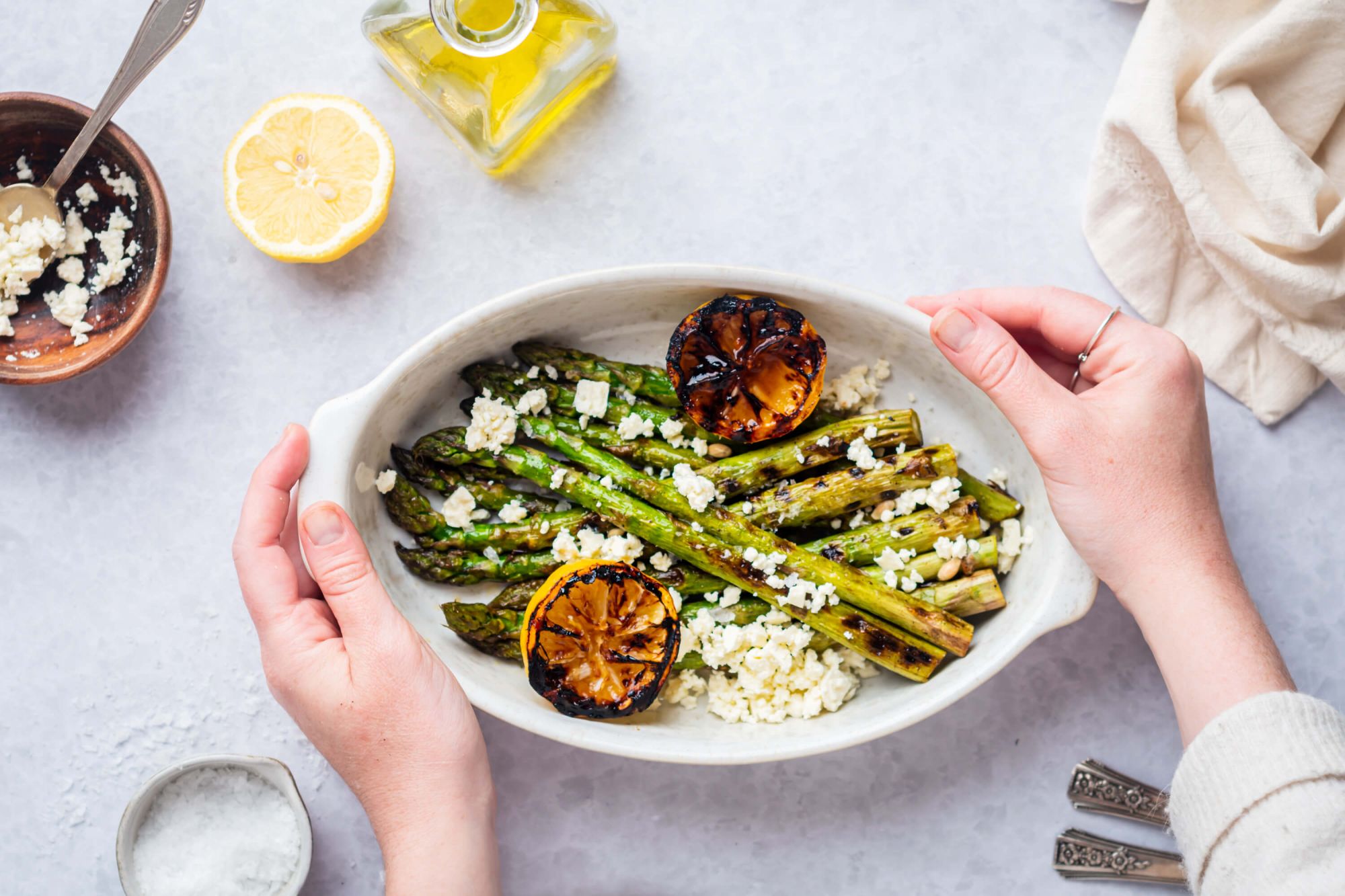 Two hands holding a plate of grilled asparagus with grilled lemon halves and crumbled feta cheese on top.