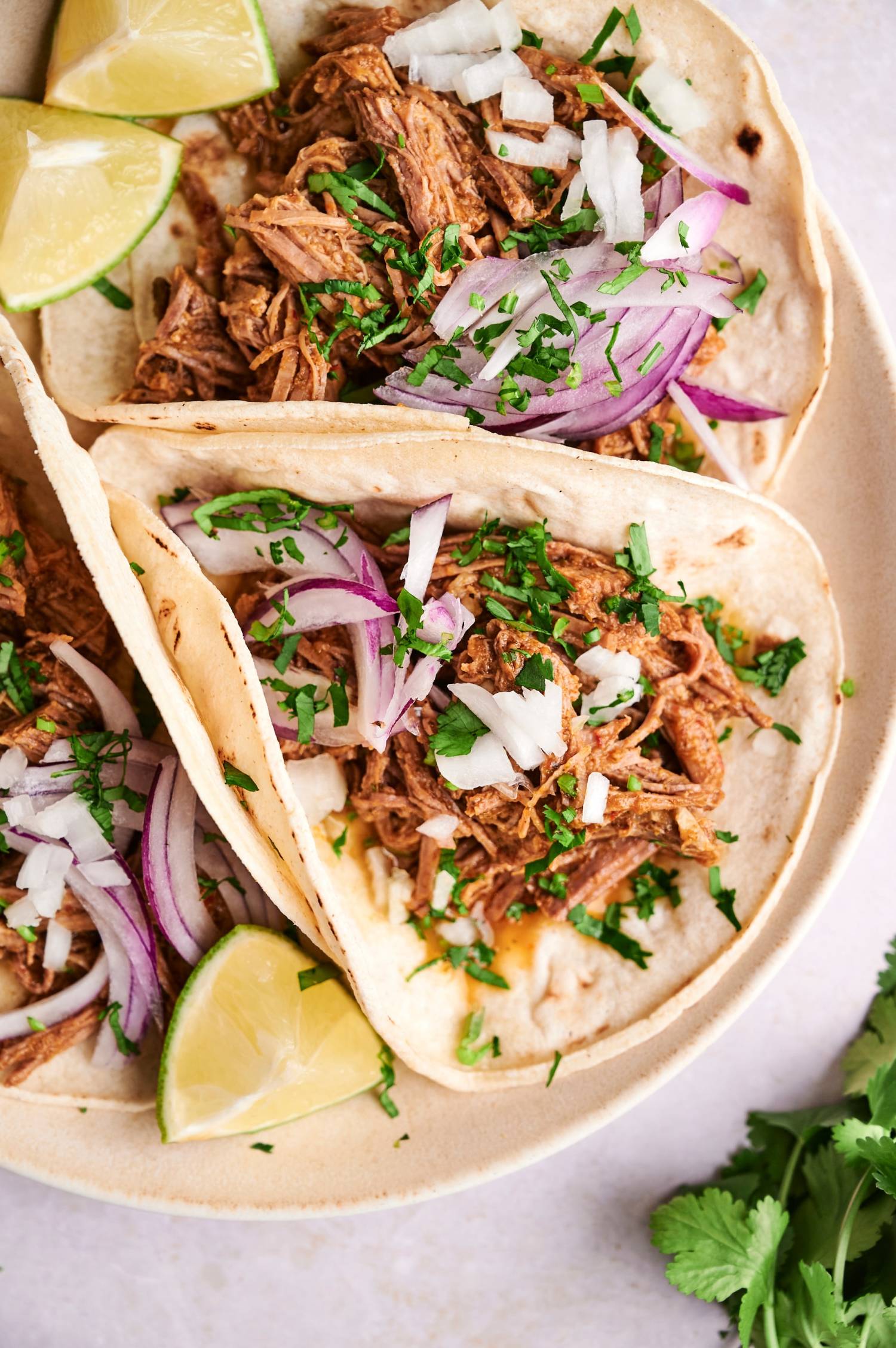 Barbacoa tacos with shredded beef, onions, cilantro, lime, and corn tortillas on a white plate.