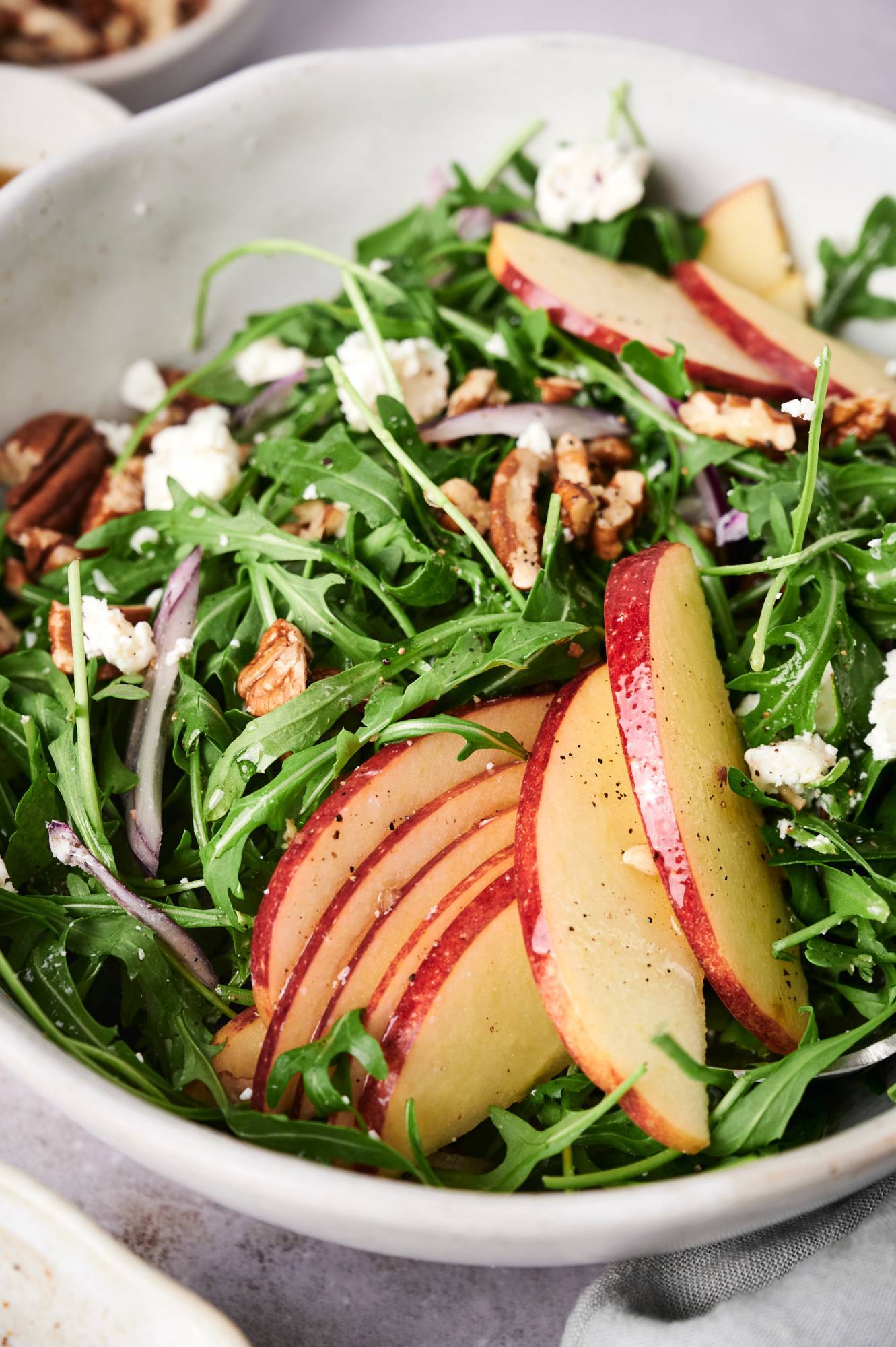 Arugula goat cheese salad with apples, pecans, and red onion served in a white bowl.