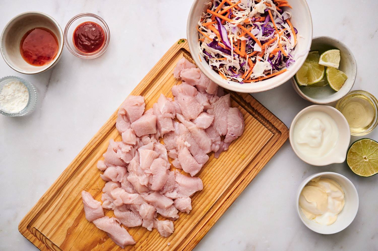 Ingredients for bang bang chicken tacos including sliced chicken breast, mayo, yogurt, Sriracha, limes, cabbage slaw, and tortillas.