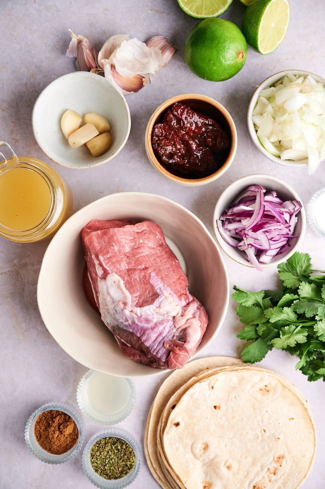 Ingredients for homemade beef barbacoa including beef roast, spices, garlic, onions, cilantro, chipotle peppers, and toritllas.