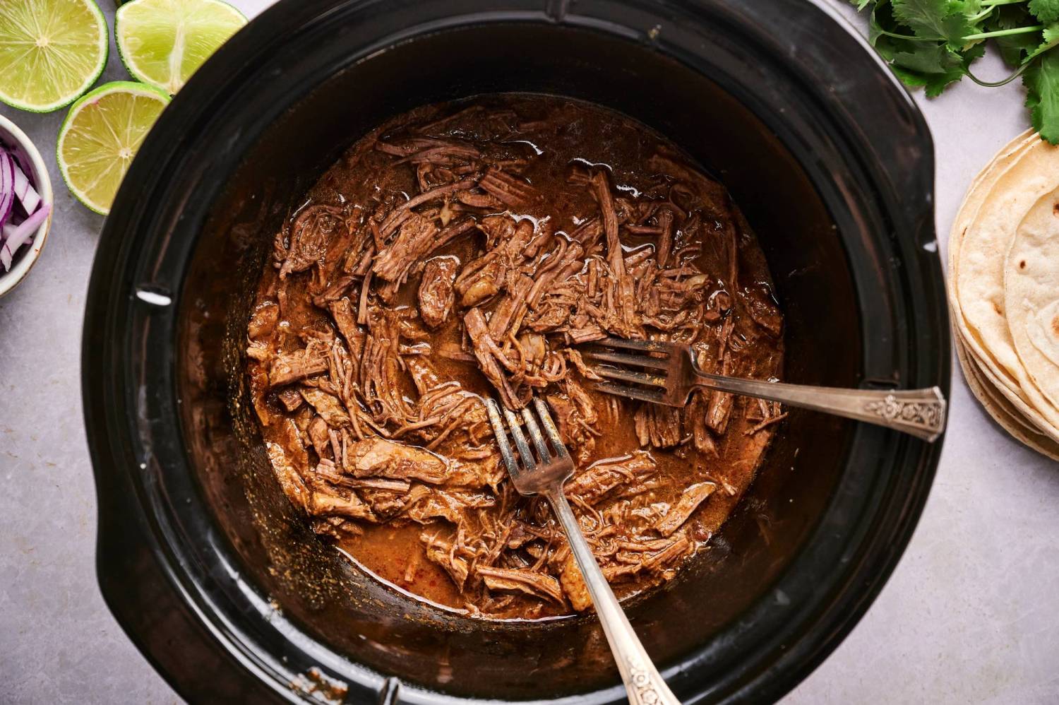 Shredded beef barbacoa in a slow cooker with chipotle chili sauce and two forks.