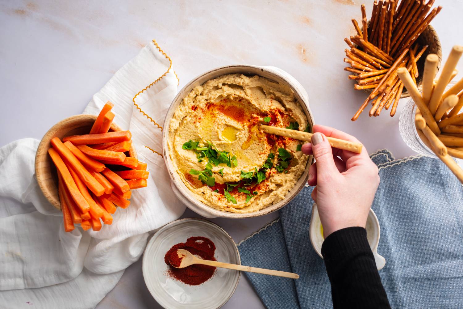 Homemade hummus in a bowl with carrots, pretzels, breadsticks, and paprika on the side.