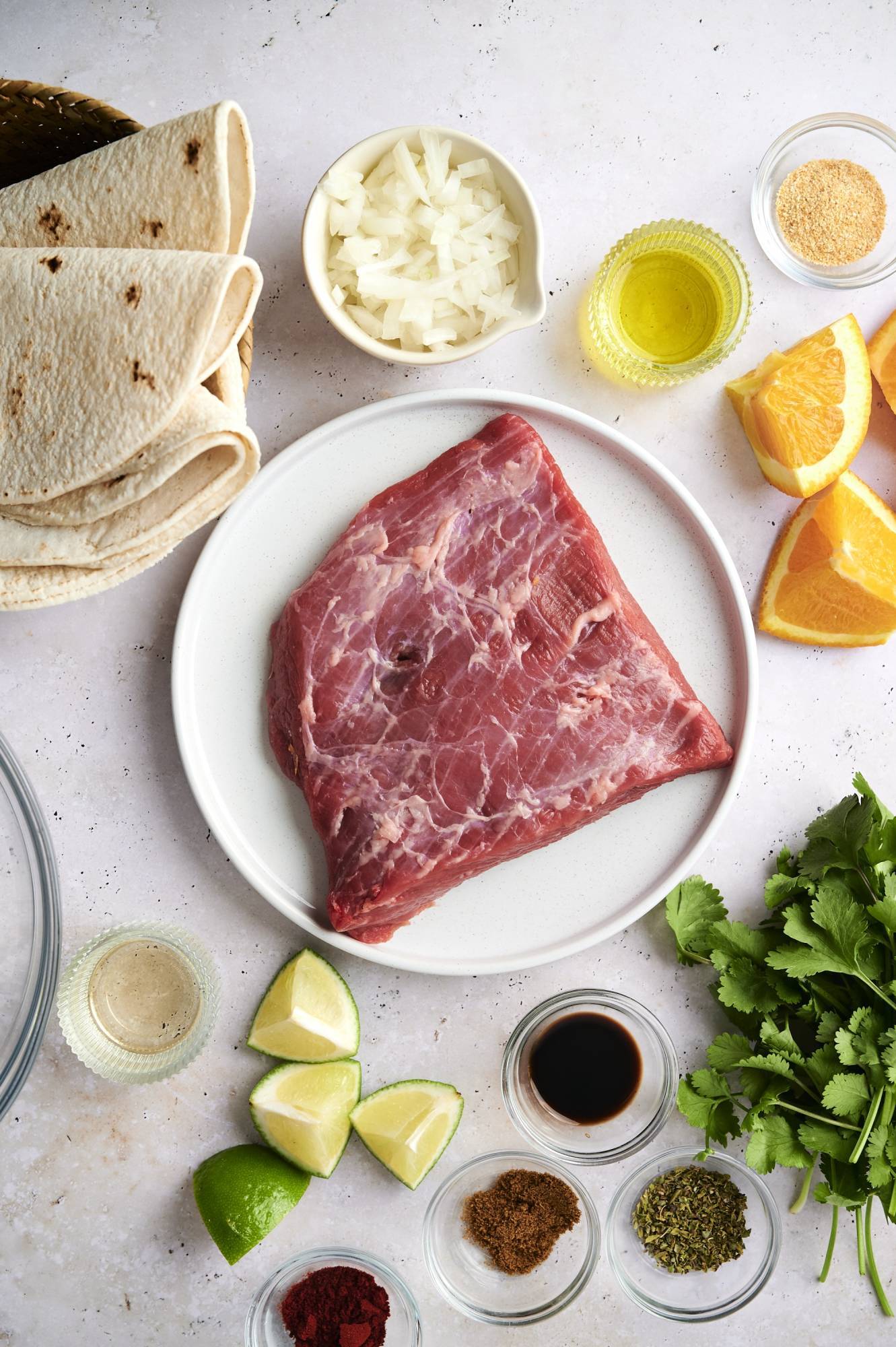 Ingredients for carne asada street tacos including flank steak, limes, oranges, cilantro, onion, spices, and tortillas.