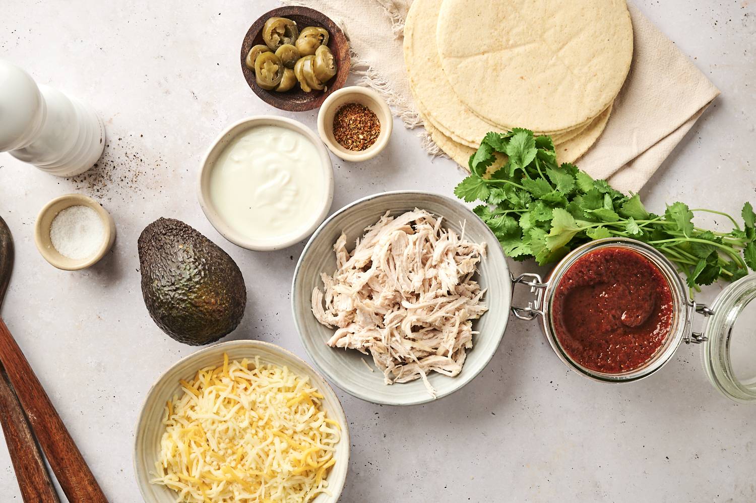 A spread of chicken enchilada ingredients, including shredded chicken, tortillas, cheese, avocado, sour cream, red enchilada sauce, jalapeños, and fresh cilantro.