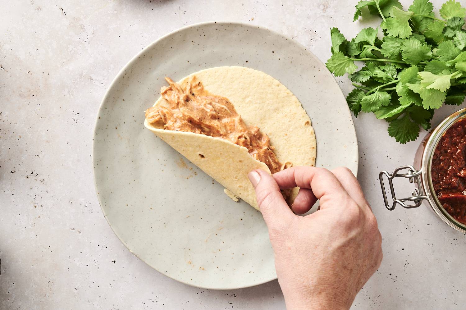 A hand folding a tortilla filled with creamy shredded chicken, ready to assemble chicken enchiladas.