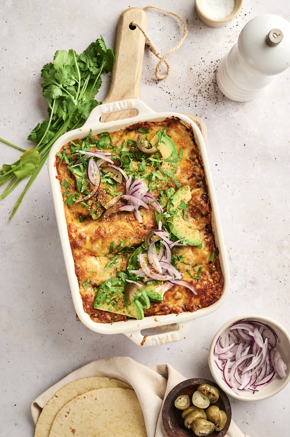 A freshly baked casserole dish of cheesy chicken enchiladas topped with avocado slices, red onion, and jalapeños, garnished with cilantro.