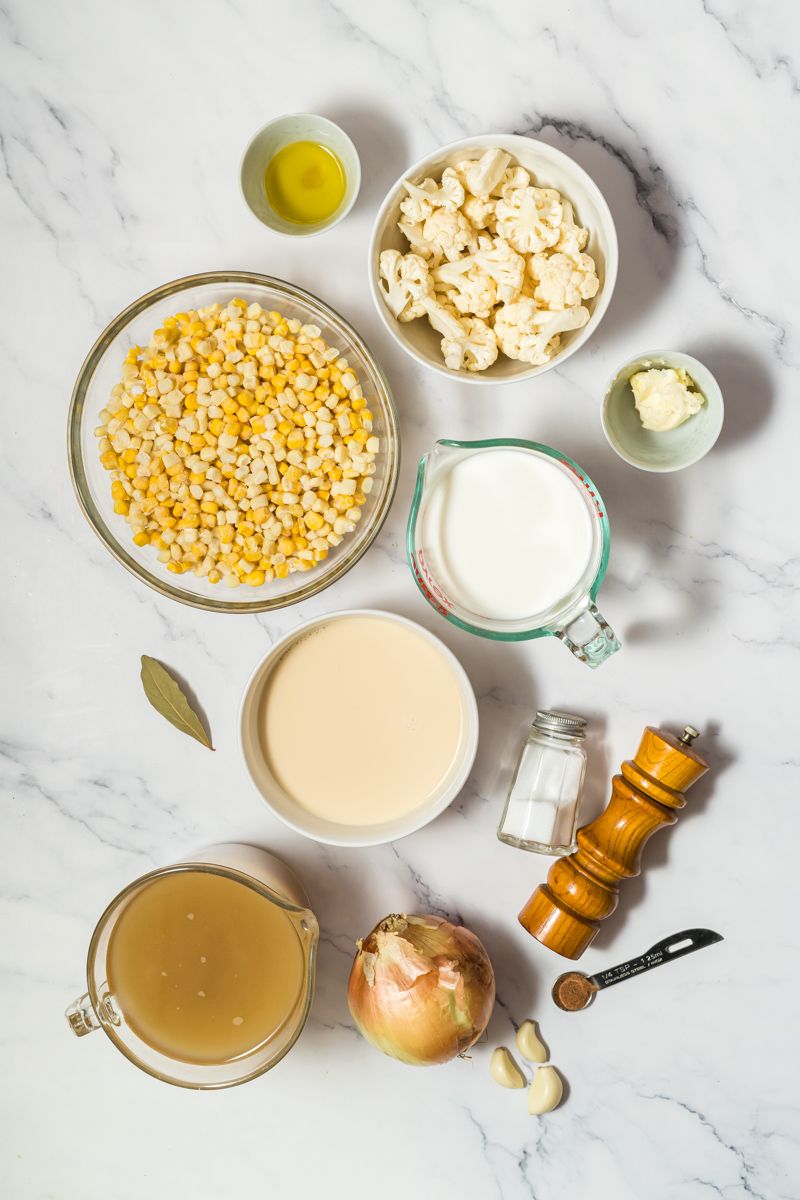 Ingredients for corn chowder including fresh corn, cauliflower, evaporated milk, whole milk, onion, and garlic on a marble board.