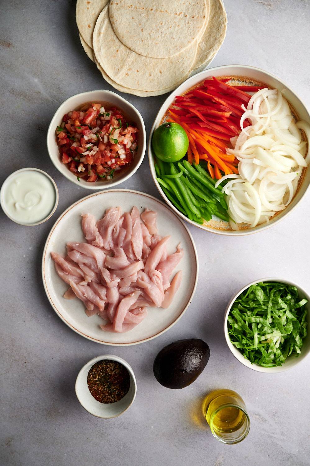 Ingredients for fajita tacos including chicken breast, bell peppers, onions, fajita seasoning, corn tortillas, limes, and toppings.